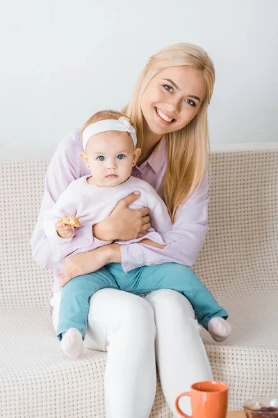 Young woman sitting on sofa with small daughter holding cookie — Stock Photo