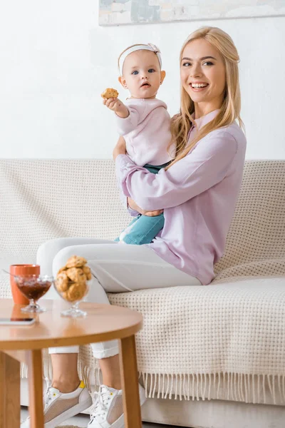 Giovane donna seduta sul divano con piccola figlia che tiene il biscotto — Foto stock