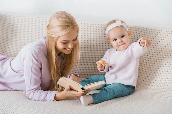 Young happy mother laying on sofa and reading book to small daughter while toddler holding cookie — Stock Photo