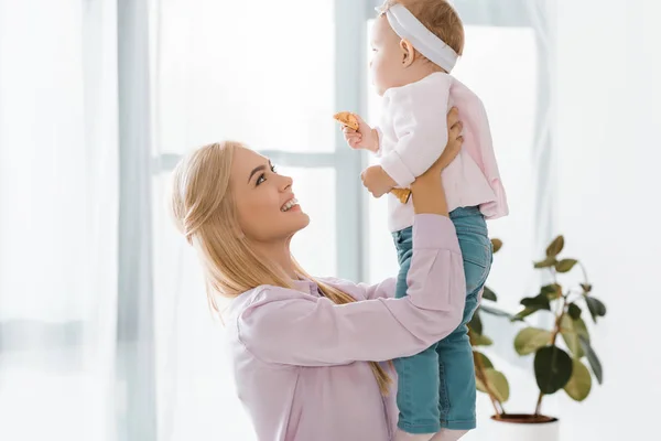 Молодая улыбающаяся мать держит малышку дочь с печеньем в руке — стоковое фото