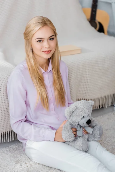 Jolie femme assise sur le tapis avec ours en peluche dans les mains — Photo de stock