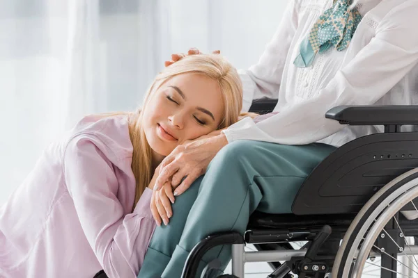 Молодая женщина с закрытыми глазами, лежащая на коленях пожилой женщины в инвалидном кресле — стоковое фото