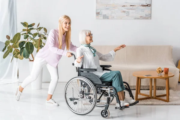 Jeune femme poussant fauteuil roulant avec heureuse femme âgée dans la maison de soins infirmiers — Photo de stock