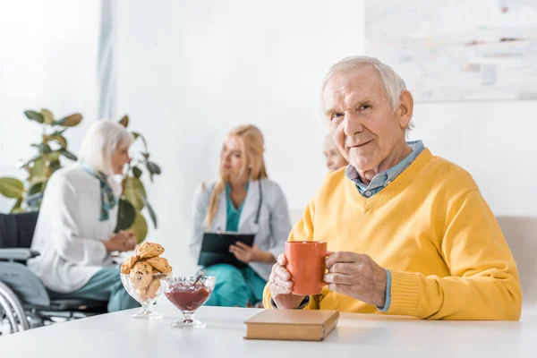 Homme âgé buvant du thé pendant que le médecin examine les femmes âgées — Photo de stock