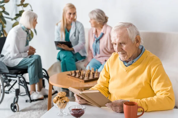 Пожилой мужчина читает книгу, а женщина-врач осматривает пожилых женщин — стоковое фото