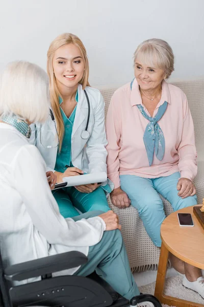 Joven enfermera sentada en el sofá y hablando con mujeres mayores - foto de stock