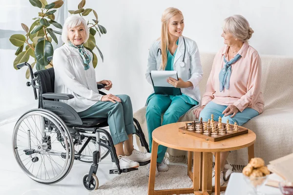 Giovane infermiera sorridente seduta sul divano e che scrive prescrizione alle donne anziane — Foto stock