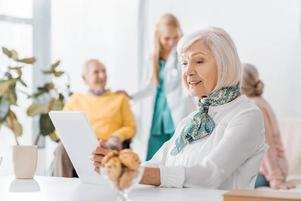 Seniorin nutzt digitales Tablet im Pflegeheim mit verschwommenen Personen im Hintergrund — Stockfoto