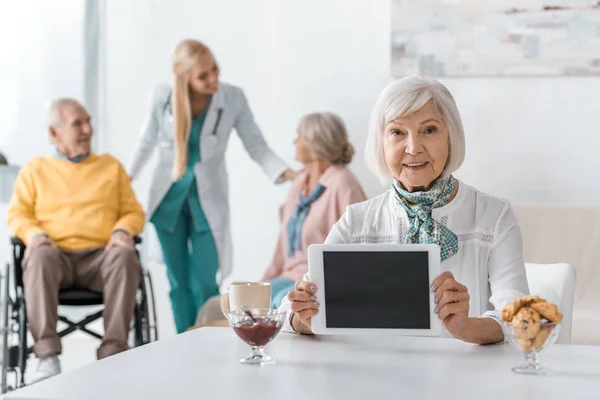 Femme âgée tenant tablette numérique à la maison de soins infirmiers avec des personnes âgées et un médecin en arrière-plan — Photo de stock