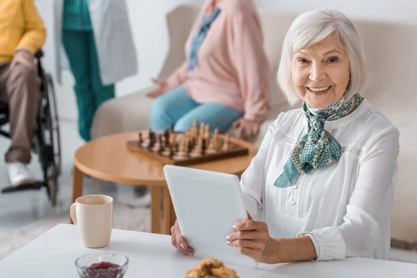 Seniorin nutzt digitales Tablet im Pflegeheim mit verschwommenen Personen im Hintergrund — Stockfoto