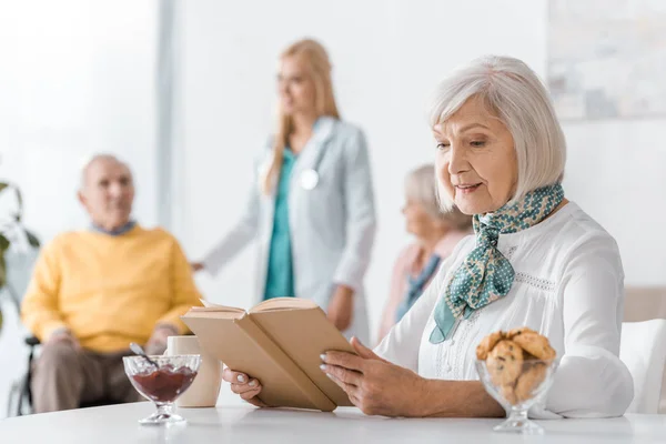 Libro di lettura donna anziana a casa di cura con persone offuscate sullo sfondo — Foto stock