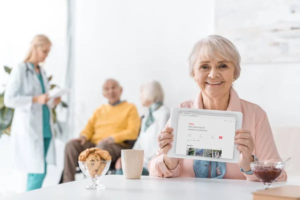 Mujer mayor mostrando aplicación airbnb en la pantalla de tableta digital en el hogar de ancianos - foto de stock