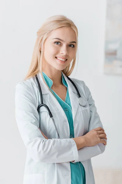 Lächelnde junge Ärztin mit Stethoskop steht mit verschränkten Armen in Klinik — Stockfoto