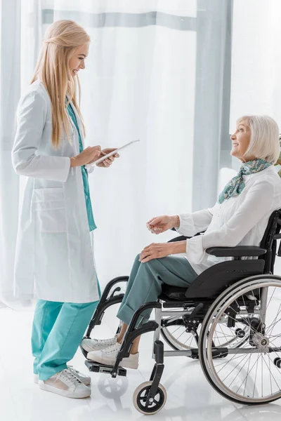 Jeune femme médecin écrit prescription sur presse-papiers pour femme âgée handicapée — Photo de stock