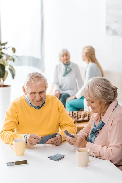 Hombre y mujer mayores jugando a las cartas en la mesa en el hogar de ancianos - foto de stock