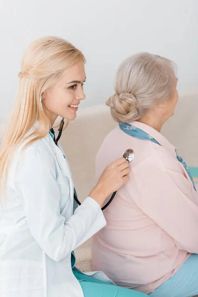 Joven sonriente médico examinando con estetoscopio la espalda de la mujer mayor - foto de stock