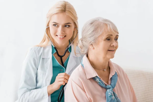 Молодая улыбающаяся женщина-врач осматривает со стетоскопом пожилую женщину — стоковое фото