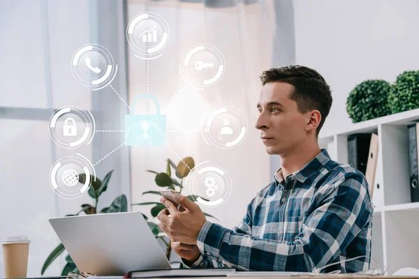 Vista lateral del hombre de negocios con teléfono inteligente sentado en el lugar de trabajo con el ordenador portátil en la oficina, concepto de seguridad cibernética - foto de stock