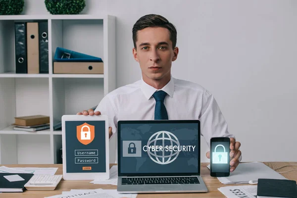 Retrato del hombre de negocios que muestra el ordenador portátil, tableta y teléfono inteligente con signos de seguridad cibernética en las pantallas en el lugar de trabajo en la oficina — Stock Photo