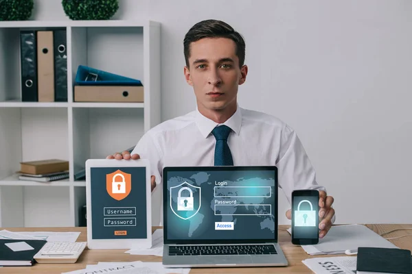 Porträt eines Geschäftsmannes, der Laptop, Tablet und Smartphone mit Cyber-Sicherheitsschildern auf Bildschirmen am Arbeitsplatz im Büro zeigt — Stockfoto