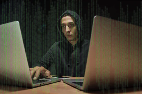 Retrato de hacker en sudadera con capucha negra usando computadoras portátiles en la mesa con teléfono inteligente, concepto de seguridad cibernética - foto de stock