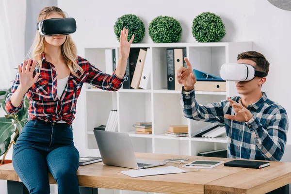 Jóvenes empresarios con auriculares de realidad virtual gesticulando en el lugar de trabajo en la oficina - foto de stock