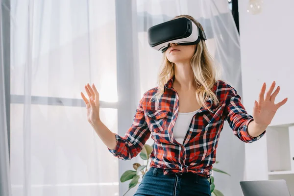 Mujer joven con auriculares de realidad virtual haciendo gestos en la habitación - foto de stock