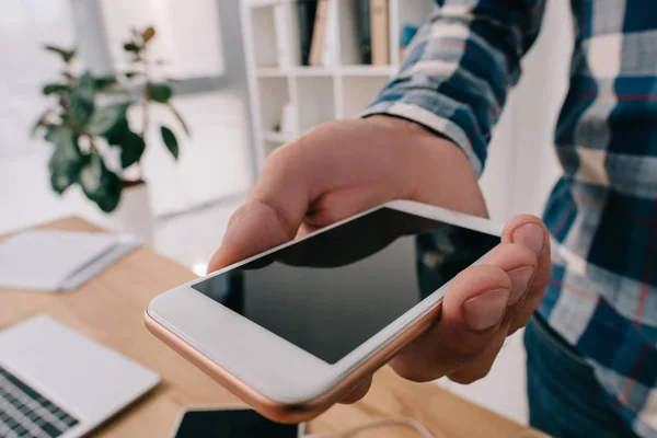 Vista parcial del hombre sosteniendo el teléfono inteligente con pantalla en blanco en la mesa con el ordenador portátil - foto de stock