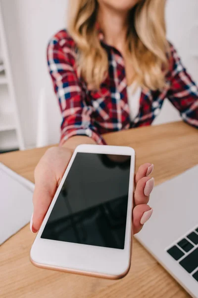 Vue partielle de la femme tenant smartphone avec écran blanc sur le lieu de travail avec ordinateur portable — Photo de stock