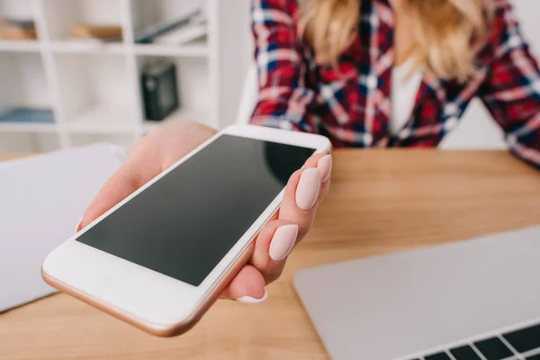 Vue partielle de la femme tenant smartphone avec écran blanc sur le lieu de travail avec ordinateur portable — Photo de stock