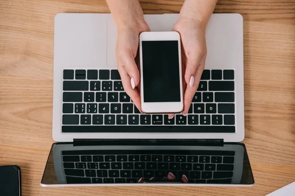 Обрезанный снимок женщины, держащей смартфон в руках на столе с ноутбуком — стоковое фото