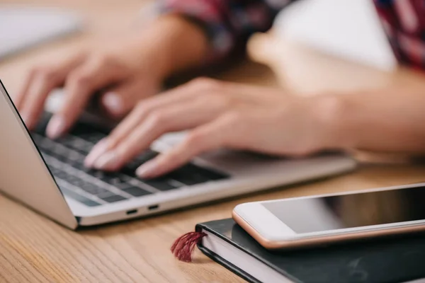 Partial view of woman typing on laptop at tabletop with smartphone and notebook — Stock Photo