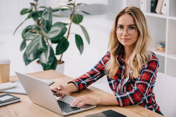 Mulher sorrindo olhando para a câmera ao usar smartphone e laptop em uma vez no local de trabalho — Fotografia de Stock