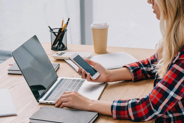 Vista parcial de la mujer de negocios utilizando el teléfono inteligente y el ordenador portátil en el lugar de trabajo con papeles en la oficina — Stock Photo