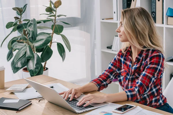 Mujer de negocios pensativa mirando hacia otro lado mientras se utiliza el ordenador portátil en el lugar de trabajo con papeles - foto de stock