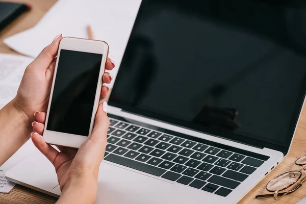 Plan recadré de femme d'affaires tenant smartphone avec écran blanc sur le lieu de travail avec ordinateur portable — Photo de stock