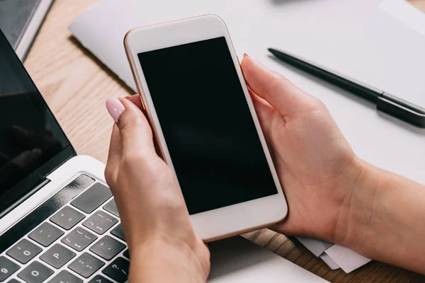 Recortado disparo de la mujer de negocios celebración de teléfono inteligente en el lugar de trabajo con el ordenador portátil - foto de stock