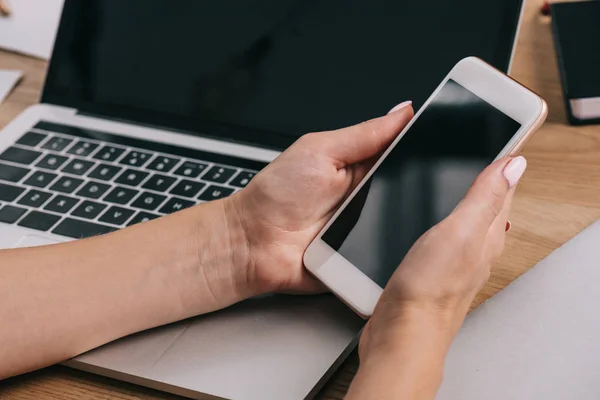 Tiro recortado de empresária segurando smartphone com tela em branco no local de trabalho com laptop — Fotografia de Stock