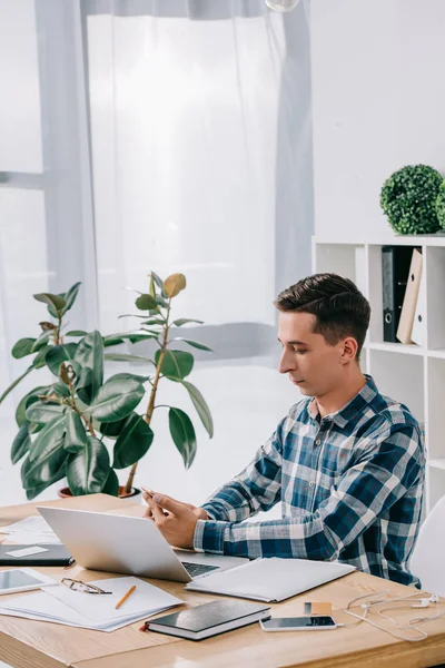 Visão lateral do empresário usando smartphone no local de trabalho com laptop no escritório e notebook — Fotografia de Stock