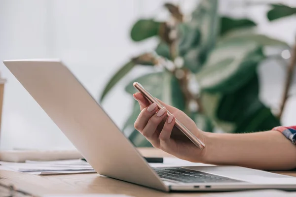 Tiro cortado de mulher de negócios com smartphone no local de trabalho com laptop — Fotografia de Stock