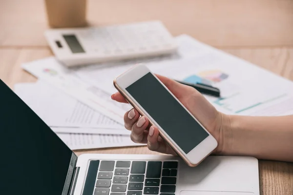 Recortado de la mujer de negocios sosteniendo teléfono inteligente con pantalla en blanco en el lugar de trabajo con el ordenador portátil — Stock Photo