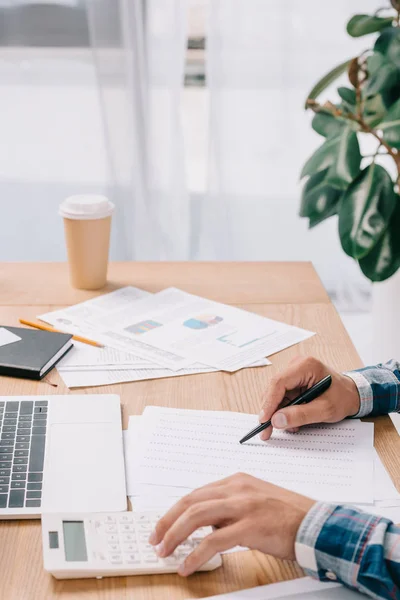 Teilansicht eines Geschäftsmannes, der am Arbeitsplatz mit Dokumenten und Laptop Berechnungen vornimmt — Stockfoto