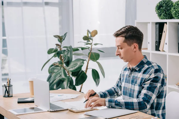 Visão lateral do empresário focado fazendo cálculos no local de trabalho com papéis e laptop no escritório — Fotografia de Stock