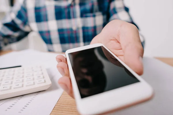 Recortado disparo de hombre de negocios mostrando teléfono inteligente con pantalla en blanco en el lugar de trabajo con papeles y calculadora - foto de stock