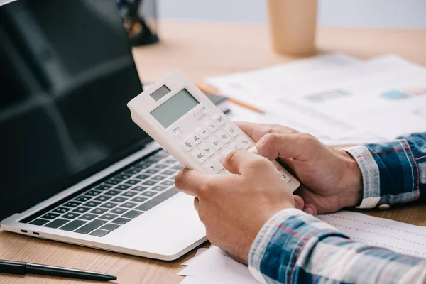 Recortado tiro de hombre de negocios con calculadora de trabajo en el lugar de trabajo con documentos y portátil - foto de stock