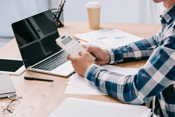 Recortado tiro de hombre de negocios con calculadora de trabajo en el lugar de trabajo con documentos y portátil - foto de stock