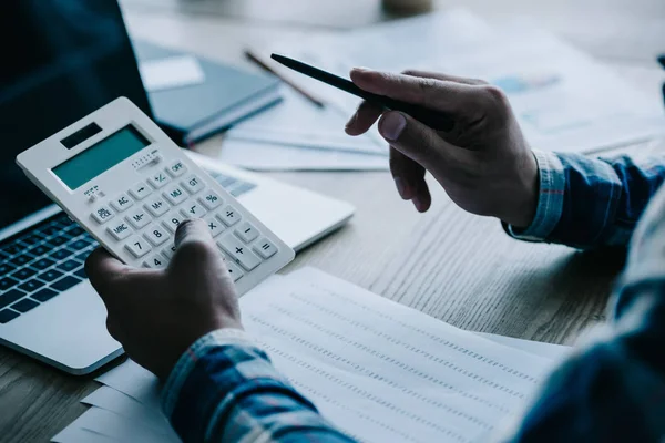 Обрізаний знімок бізнесмена з калькулятором, що працює на робочому місці з документами та ноутбуком — стокове фото