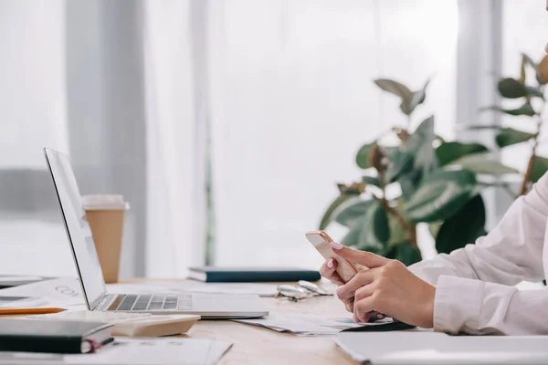 Vista parcial de la mujer de negocios con teléfono inteligente en el lugar de trabajo con documentos y portátil en la oficina - foto de stock