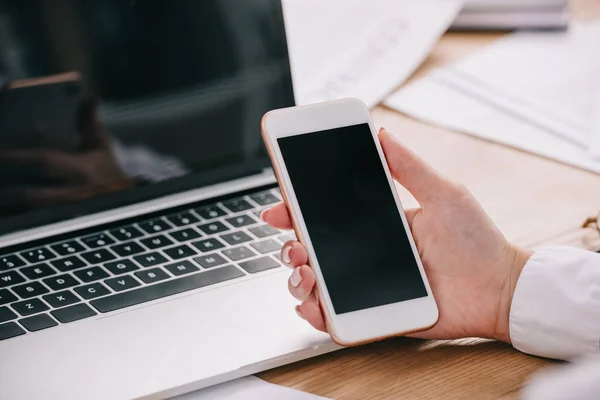 Обрезанный снимок предпринимательницы, держащей смартфон на рабочем столе с ноутбуком — стоковое фото