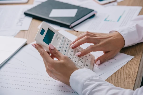 Recortado disparo de mujer de negocios haciendo cálculos en la calculadora en el lugar de trabajo con documentos - foto de stock